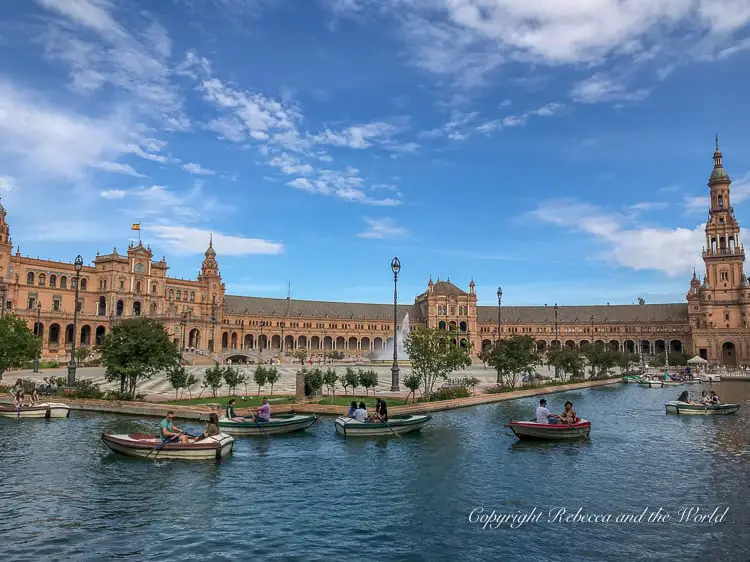 A large plaza with a central building and a moat where people are rowing small boats, under a blue sky with clouds. This is the beautiful Plaza de Espana in Seville, Spain.