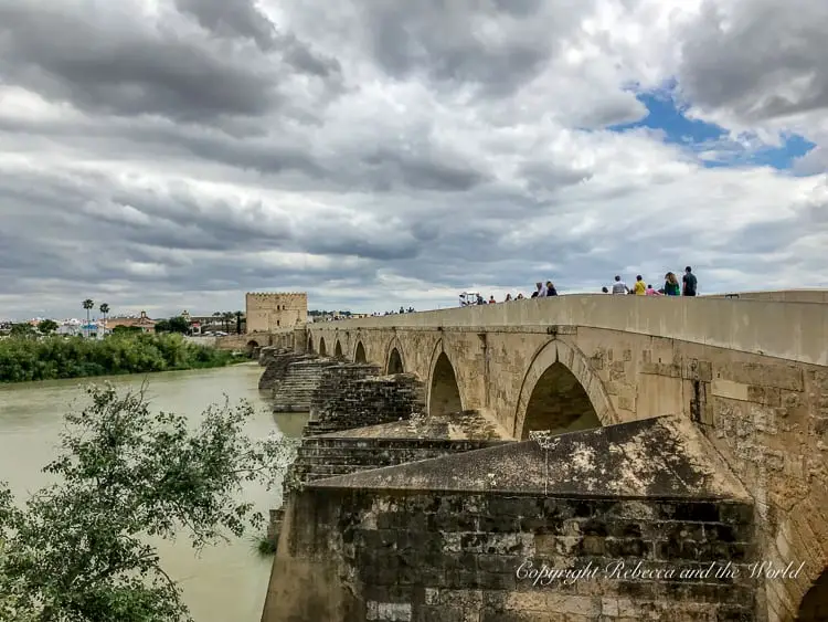 A stone medieval bridge with multiple arches crossing a river, with people visible walking across under a cloudy sky. This is the Roman Bridge in Cordoba, Spain.