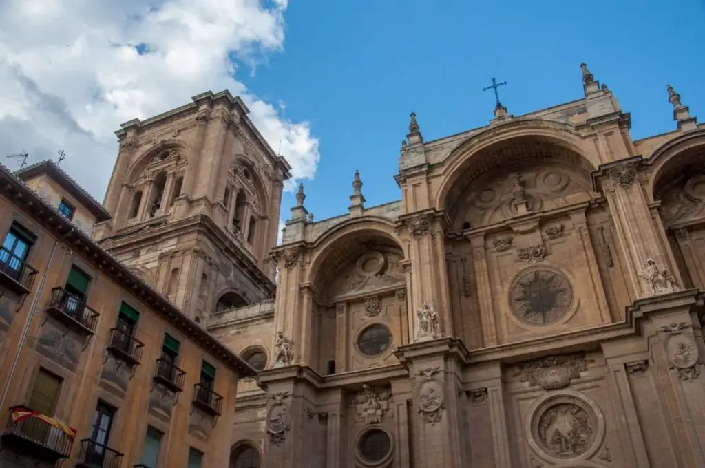 Upward view of a majestic cathedral façade showcasing Baroque architectural details with sculptures, circular windows, and two bell towers against a clear blue sky. This is the Granada Cathedral.