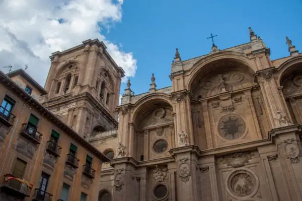 Upward view of a majestic cathedral façade showcasing Baroque architectural details with sculptures, circular windows, and two bell towers against a clear blue sky. This is the Granada Cathedral.