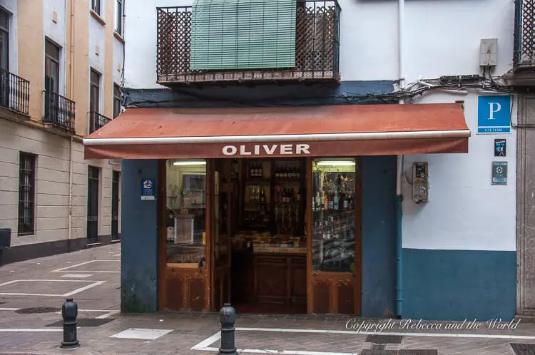 A street corner with a vintage shop named 'OLIVER,' featuring a wooden storefront and an awning. The shop is situated at the intersection of two pedestrian streets with bollards. This is the oldest store in all of Spain, and is found in Granada Andalucia.