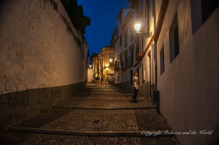 An evening view of a quaint cobblestone alleyway with white walls, illuminated by warm streetlights and a blue twilight sky, with a few pedestrians walking along the path. This is the Albaicin, a popular area to stay and visit in Granada, Spain. I recommend staying here if you have 2 days in Granada, as it's close to the main attractions.