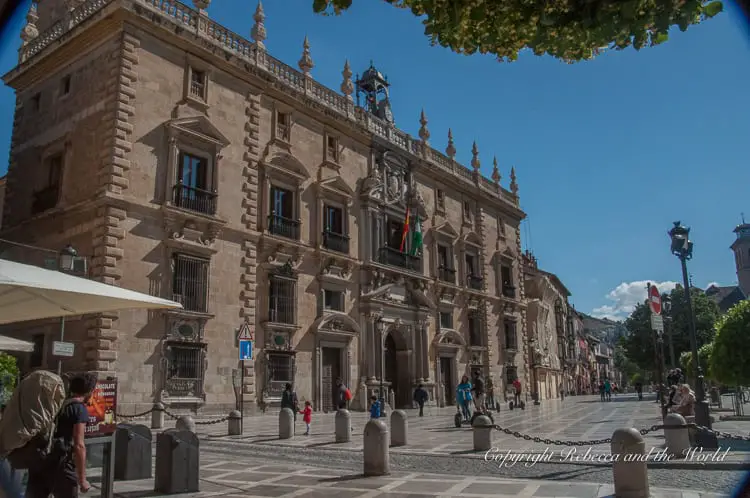 A grand historical building in Plaza Nueva, Granada, with intricate façade details and multiple arched windows, under a clear blue sky. People are walking by and sitting on the bollards on the wide pedestrian path in front of the building. 2 days in Granada means beautiful sights like this.