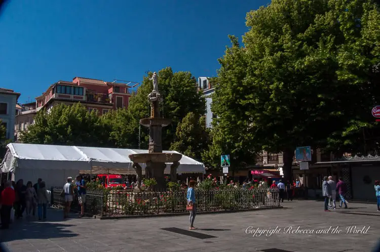 A bustling square with a classic stone fountain at the center surrounded by a metal fence, shaded by trees, and lined with market tents, with people milling around in the sunlight. This is one of the many squares in beautiful Granada, Spain.