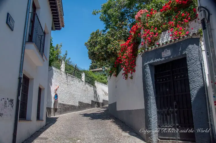 A narrow cobblestone street flanked by white walls and a house with a dark green door. Lush green plants and vibrant red flowers are draping over the wall on the right side under a bright sunny sky. I love wandering the streets of Granada.