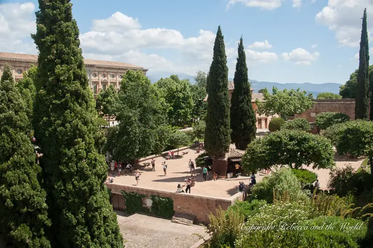 A public square surrounded by trees and historical buildings, with people walking and sitting around. The square is viewed from an elevated position, framed by tall cypress trees. This is the amazing Alhambra - one of the most amazing places to visit in Granada, Spain.