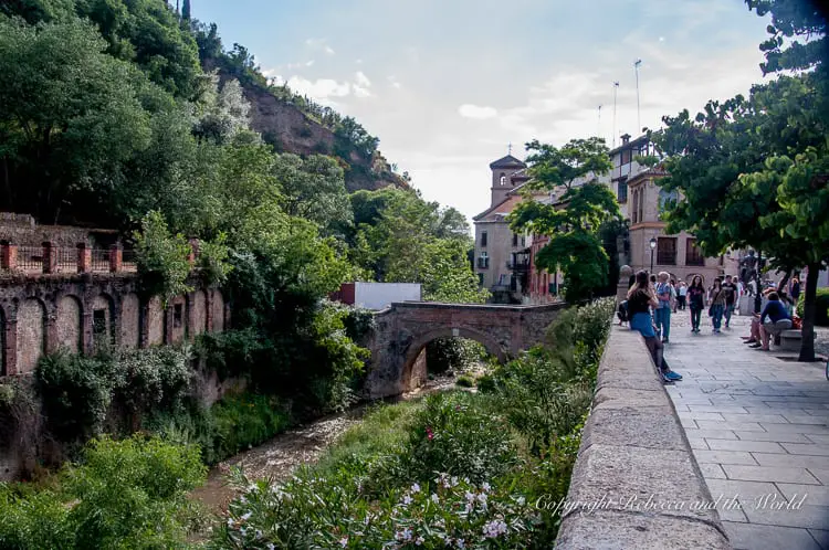 A scenic view of an ancient stone bridge crossing a river, with lush greenery and trees. People are walking along a path next to the bridge under a partly cloudy sky. This area is known as the Paseo de los Tristes in Granada, Spain.