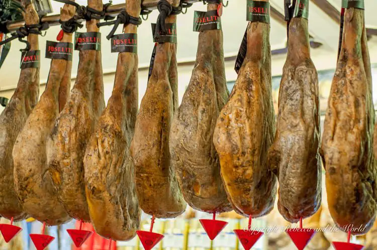 A display of hanging cured ham legs, known as 'jamón,' with black hoof ends. They are labeled with 'JOSELITO' tags, indicating a brand, and suspended from metal hooks in a market setting.