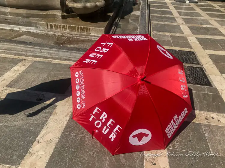A close-up of a red umbrella with the words 'FREE TOUR' printed on it, along with the logo of a walking figure and 'WALK IN GRANADA' text, indicating a tour service point. A free walking tour is a great way to start off your Granada itinerary.