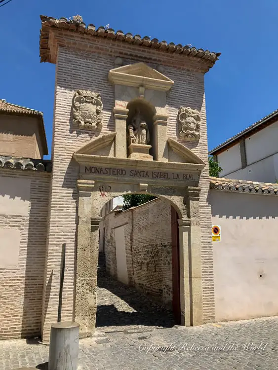 Entrance to the 'Monasterio Santa Isabel La Real' with a stone archway bearing the name and decorative elements, including a small statue and crests, leading into a shaded alley. One of the most unique things to do in Granada is visit the Santa Isabel Convent to buy sweets from the cloistered nuns.