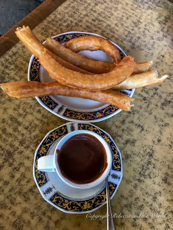 A traditional Spanish snack consisting of churros, long fried dough pastries, served on a white and blue ceramic plate with a matching cup of thick hot chocolate for dipping.