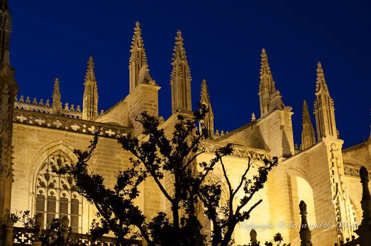 The Seville Cathedral illuminated at night, highlighting its impressive Gothic features and flying buttresses against the night sky. Barrio Santa Cruz is a great area to explore at night, when the Seville Cathedral is lit up and street performers entertain visitors.