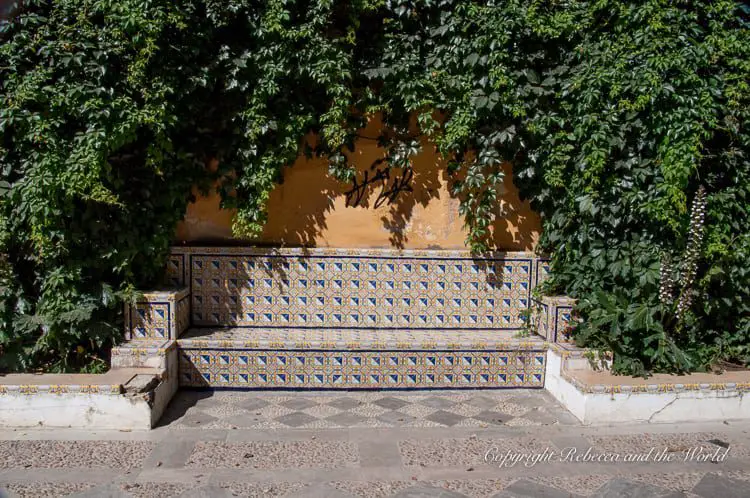 A quaint bench in Seville, Spain, built against a peach-colored wall, adorned with intricate, blue and yellow patterned tiles. The bench is partially obscured by lush green foliage from an overhanging tree. The scene appears peaceful and secluded.