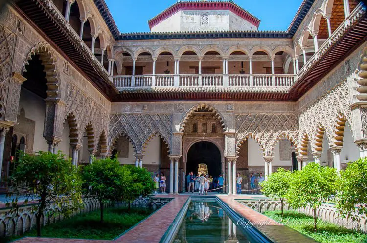 The grand courtyard of the Real Alcazar in Seville, Spain, within an ornate, historical building, characterized by its detailed Moorish architecture. A narrow, rectangular pool lined with greenery reflects the intricate designs of the surrounding archways, columns, and the second-story balustrades. Tourists are visible in the background, admiring the beauty of the space.