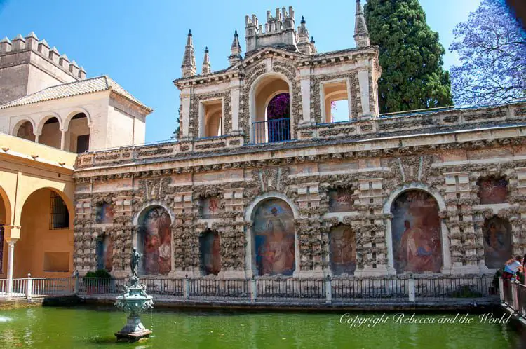 The intricate Gallery of the Grotesques in Seville, with Renaissance-era murals and elaborate stone carvings adorning the walls. A small water fountain is centered in a reflecting pool, all set against the striking architecture with arched windows and a decorative balcony. The Real Alcazar is one of the most beautiful places to see in Seville - it was even used as a filming location for Game of Thrones.