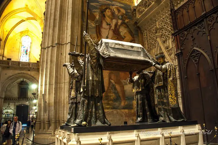 A sepulchral monument in a dimly lit cathedral, featuring four statues bearing a tomb, with detailed Gothic-style artistry. The Seville Cathedral is the final resting place of Christopher Columbus.