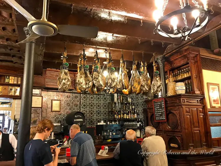 A traditional bar in Seville, where patrons are engaged in conversation. Hanging hams and an ornate wooden bar create an authentic Spanish atmosphere.