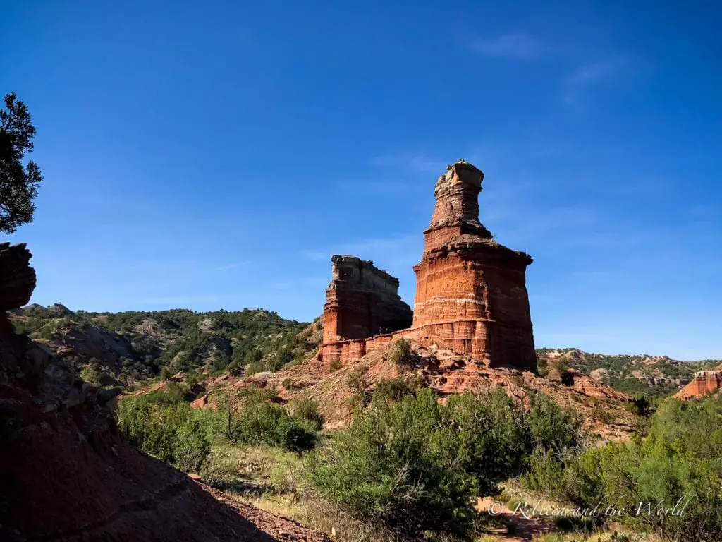 Two towering red rock formations, known as the Lighthouse structures, rising prominently above the greenery at Palo Duro Canyon State Park near Amarillo. One of the coolest things to do in Amarillo is visit nearby Palo Duro Canyon, the second largest canyon in the United States.