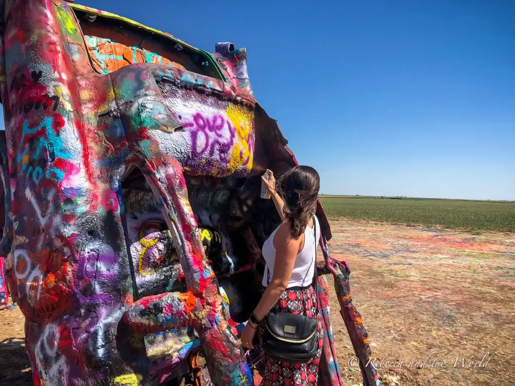 A woman in a red skirt and white singlet top sprays paint on an old Cadillac in the desert in Amarillo, Texas