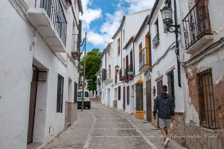 A street scene in Ronda with a person walking down a narrow street lined with white-washed houses and wrought-iron balconies, reflecting the everyday life and architectural style of the town.