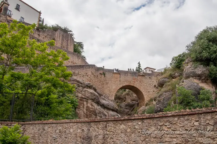 A historical arched bridge in Ronda, known as the Arab Bridge, set against a backdrop of lush greenery and ancient city walls, reflecting Ronda's rich history.