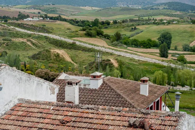 The image depicts a view over the terracotta rooftops of Ronda's buildings with traditional chimneys, with a backdrop of the rolling hills and farmlands of the Andalusian countryside, embodying the rural charm of southern Spain.