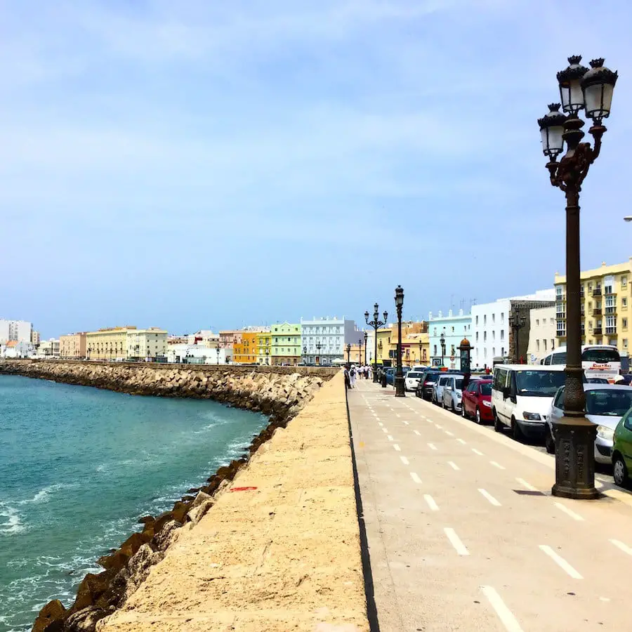 Seaside promenade in Cádiz with a line of ornate lampposts, a protective sea wall, colorful buildings in the background, and parked cars on the street under a clear blue sky.