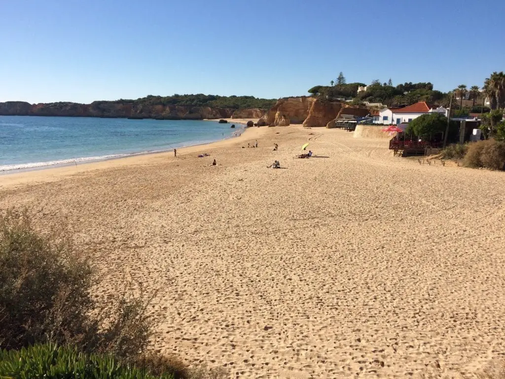 A serene and spacious sandy beach on the Algarve in Portugal, with a few people enjoying the sun, backed by green hills and a clear blue sky.