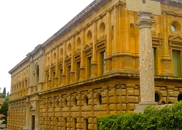 The grand facade of the Renaissance Palace of Carlos V in Granada, featuring rusticated stone walls and a series of circular medallions set above a portico of classical columns.