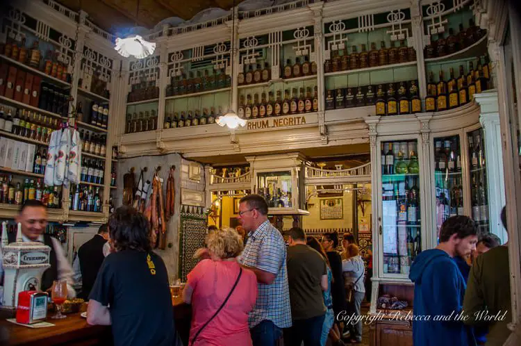 The interior of bustling El Rinconcillo tapas bar in Seville, Spain, with its vintage ambiance. The back wall is lined with shelves packed with a variety of bottles, reaching up to a high ceiling. Customers stand at the bar, engaged in conversation, while a bartender, smiling and conversational, attends to them. The decor includes tiled patterns and hanging cured meats, contributing to the traditional and cozy atmosphere. The lighting is warm, casting a soft glow over the patrons and the wooden bar area.