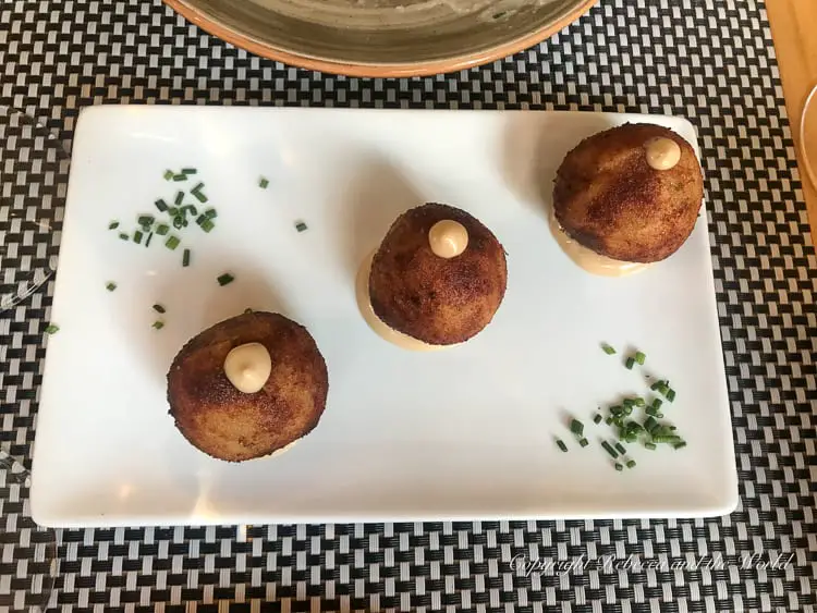 A white plate with three spherical fried croquettes, each topped with a small dollop of aioli. The plate is decorated with a scattering of chopped chives.