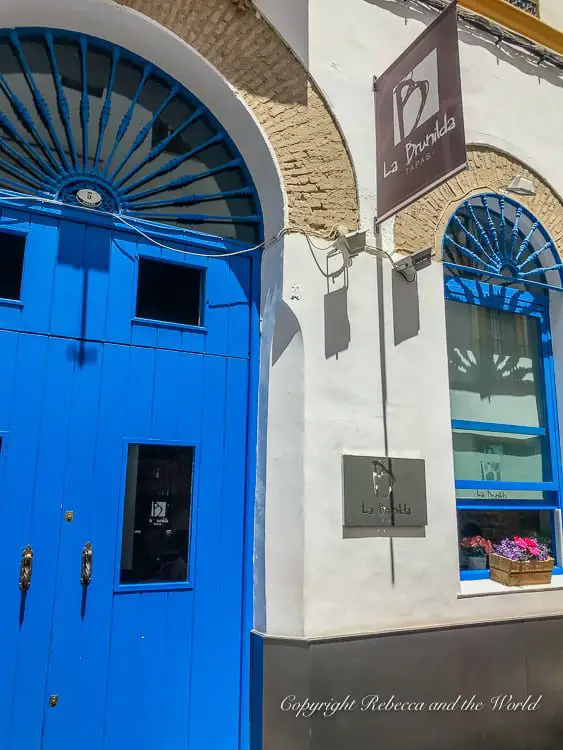 A vibrant blue double door entrance of a bar named "La Brunilda". The facade is white, and there's a sign hanging to the right displaying the bar's name and the word "Tapas." The door has ornate gold handles, and there are flower boxes with pink flowers on the window sills. This is La Brunilda, one of the most popular tapas bars in Seville, Spain.