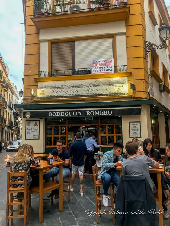 An exterior view of "Bodeguita Romero," a Seville tapas bar on a corner street. The bar has a yellow upper facade with a balcony and a window with a sign indicating an apartment for sale. Below, patrons sit at wooden tables on the sidewalk, engaged in conversation and dining.