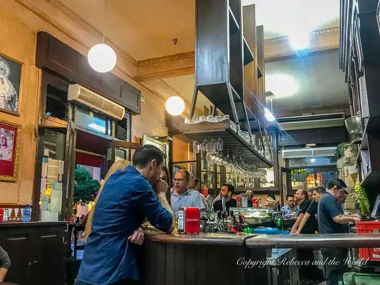 The interior of Bodegas Santa Cruz, a traditional Seville tapas bar with patrons gathered around the wooden counter. The bar is well-lit with spherical hanging lights, and the background shows a mirror reflecting the interior. People are engaged in conversation, with drinks and red napkin holders on the counter.