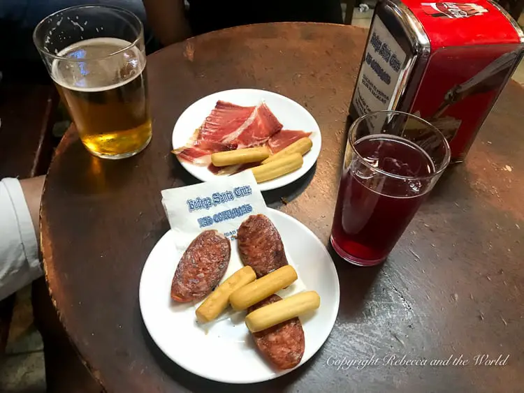 A table in a bar with a pint of amber beer and a glass of tinto de verano. There are two plates, one with slices of cured ham and breadsticks, the other with two pieces of grilled chorizo alongside more breadsticks. A napkin dispenser with the bar's name is also on the table. This is Bodega Santa Cruz, a popular tapas bar in Sevilla, Spain.