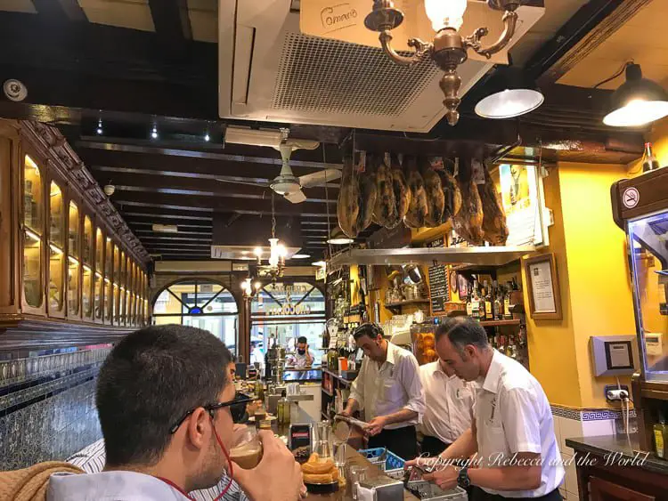 The interior of Bar El Comercio in Seville, Spain, a traditional bar with patrons seated at the counter, enjoying their drinks. Overhead hangs a selection of cured ham legs, and the bar staff is busy serving. The establishment has a warm, inviting atmosphere with wood paneling and tiled walls. It's one of the best tapas bars to try in Sevilla.