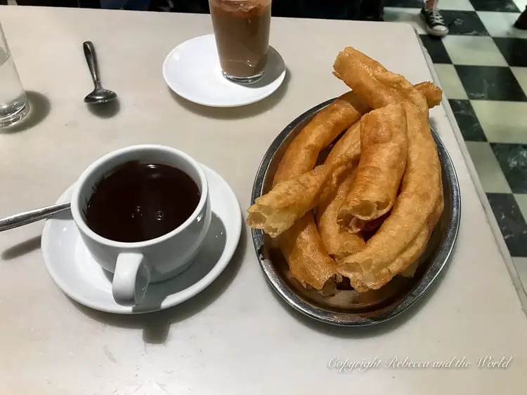 A close-up of a traditional Spanish breakfast, featuring a cup of thick hot chocolate with a plate of churros, a long, fried pastry for dipping. The table also has a glass of a milky coffee.