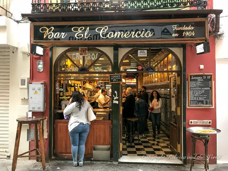 The entrance to "Bar El Comercio," established in 1904, with an ornate wooden and glass facade. Inside, patrons are visible at the bar, and a woman is standing outside, engaged in a phone conversation. The bar has a classic European style, with a checkered floor visible from the entrance.