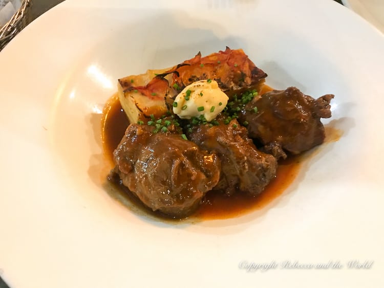 A close-up of a dish with braised beef cheeks in a rich sauce. Accompanying the meat is a roasted half potato, all served on a white plate. This is one of the tapas dishes at La Azotea in Seville, Spain.