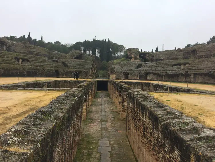 The ancient Roman amphitheater of Italica near Seville, showcasing the extensive ruins with rows of seating, an arena, and an underground passage, hinting at its historical grandeur.