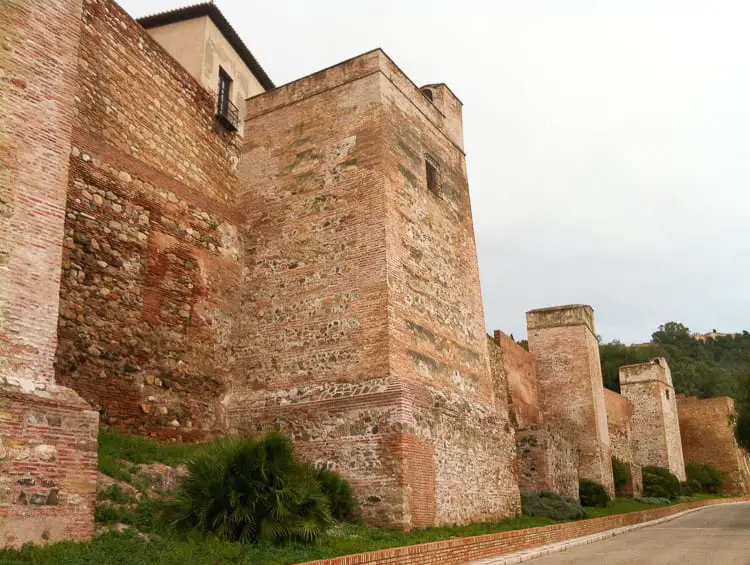 The robust, ancient walls of the Alcazaba in Málaga, showing the fortified structure’s mix of brick and stone, set against a backdrop of greenery and a cloudy sky.