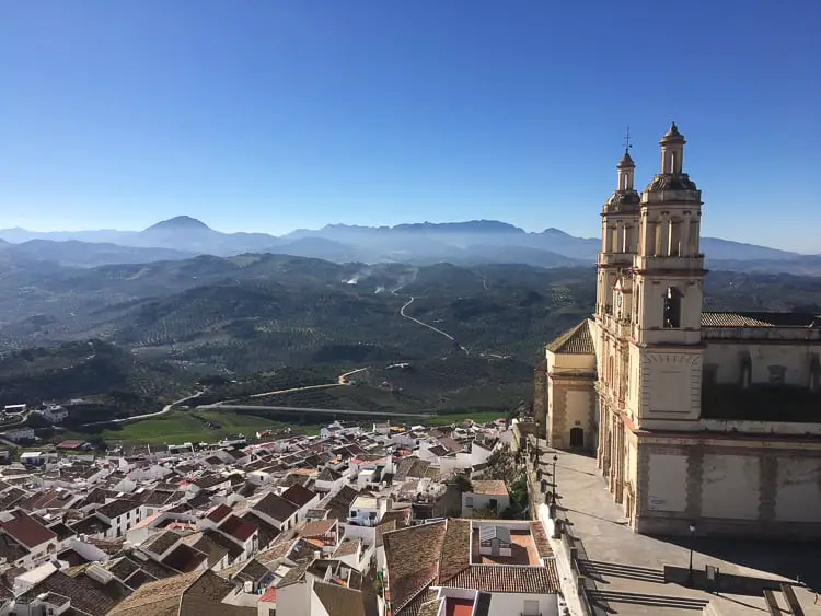 The panoramic view from the town of Olvera, showing the white houses and the Church of Nuestra Señora de la Encarnación, with the Andalusian countryside and mountains stretching into the distance.