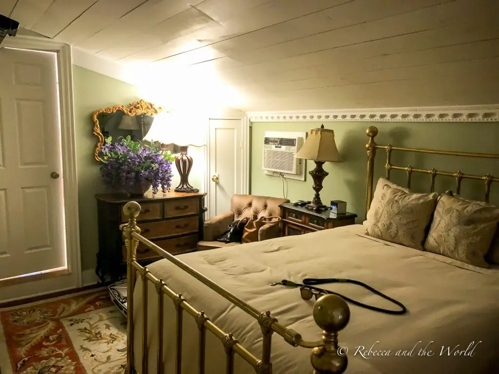 A quaint and cozy bedroom with a brass bed frame, antique furniture, a bouquet of flowers on a dresser, and a vintage feel. There are plenty of great places to stay in New Braunfels, including lovely B&Bs like Zink Haus.