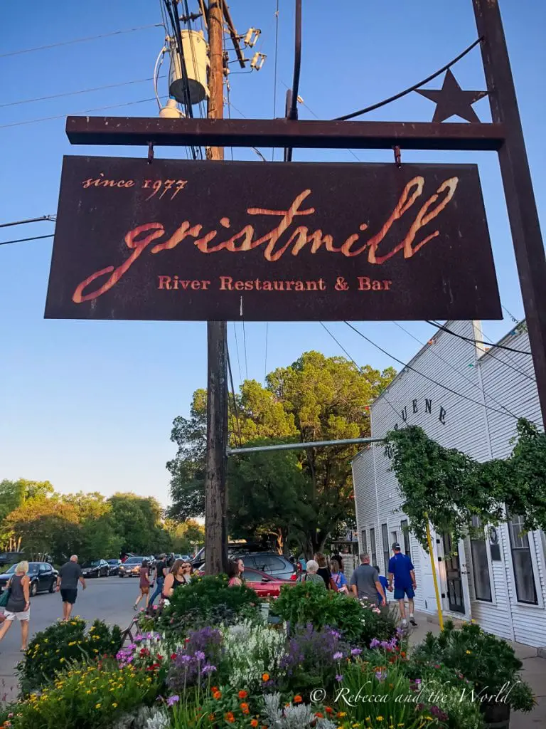 The sign for "Gristmill River Restaurant & Bar" with a backdrop of a crowded street in the evening, vibrant flower beds, and the white exterior of a building. Wondering where to eat in New Braunfels? Start at the iconic Gristmill restaurant.