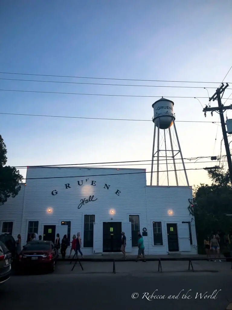 A white building with "GRUENE HALL" in bold letters, under a blue sky at dusk, with a water tower in the background and people milling about. Seeing a show at the Gruene Hall should be top of your list of things to do in New Braunfels.