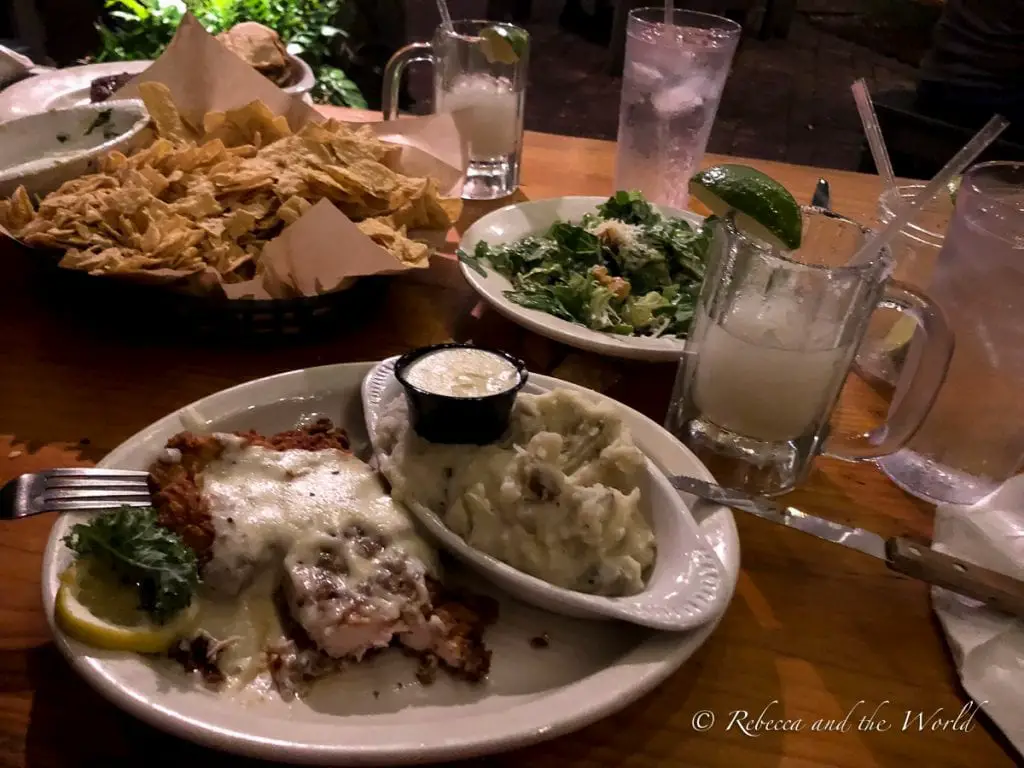 A table in a restaurant with a plate of chicken fried steak and mashed potatoes, a salad, tortilla chips and refreshing drinks including a margarita. The Gristmill in New Braunfels is a restaurant known for its comfort food like fried chicken steak.