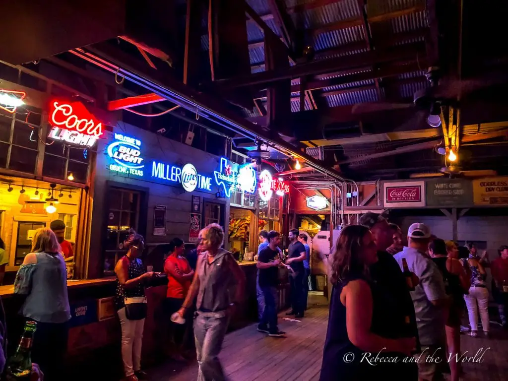 Inside a lively bar with neon signs for beers, patrons engaging in conversation, and a wooden interior illuminated by warm lighting. Memorabilia dating back to the 30s line the walls of Gruene Hall near New Braunfels, TX.