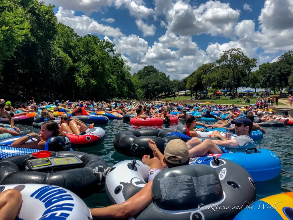 The Comal River in New Braunfels Texas crowded with people on inflatable tubes, enjoying a sunny day of floating and relaxation under a partly cloudy sky. The absolute best thing to do in New Braunfels is float the river!