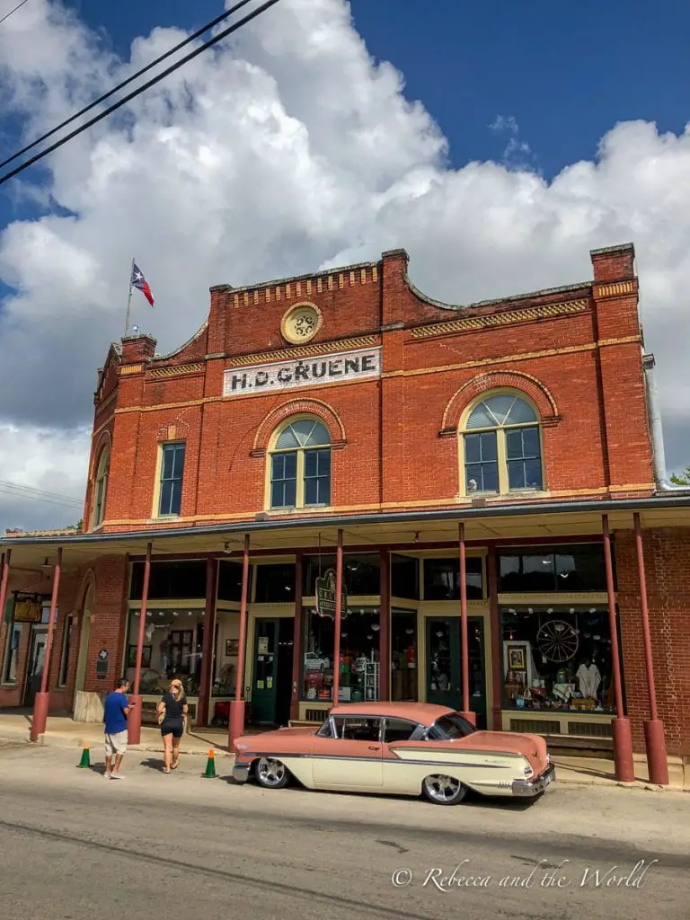 A historic brick building with "H.D. GRUENE" on the facade under a clear sky, featuring a classic two-tone car parked in front and people walking by. One of the best things to do in New Braunfels, Texas, is wander the historic Gruene area, where some original buildings still stand.