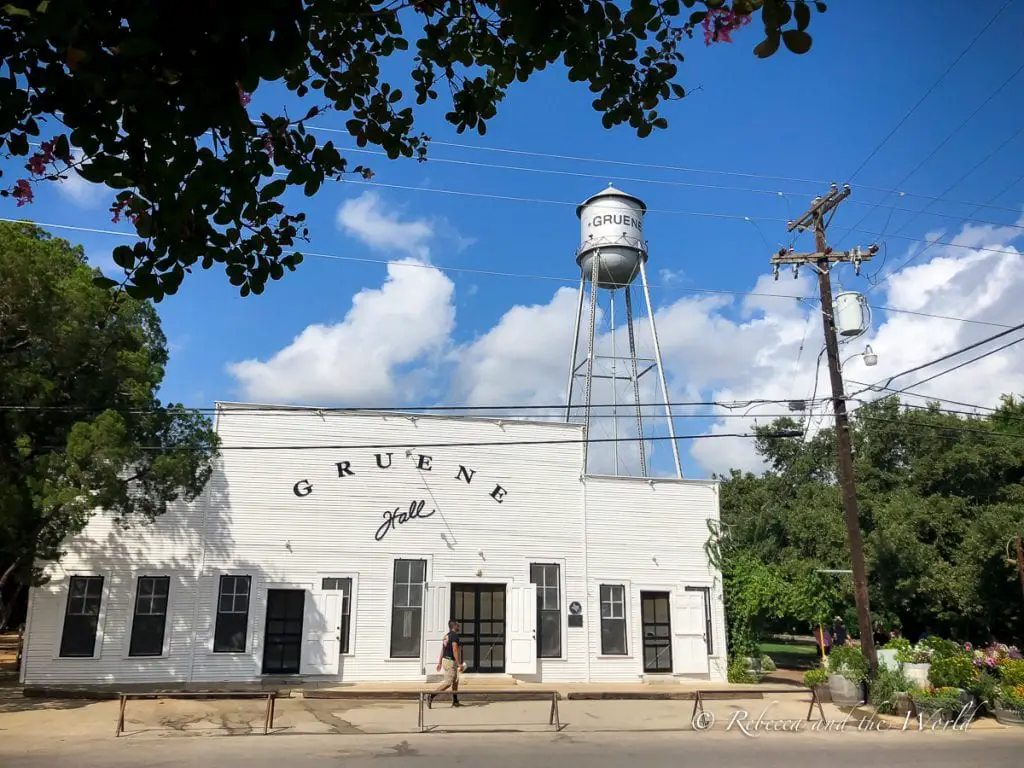 The exterior of GRUENE Hall during the day, with the water tower visible in the background and surrounded by lush trees under a partly cloudy sky. The Gruene water tower behind Gruene Hall is one of the most photographed places in New Braunfels.